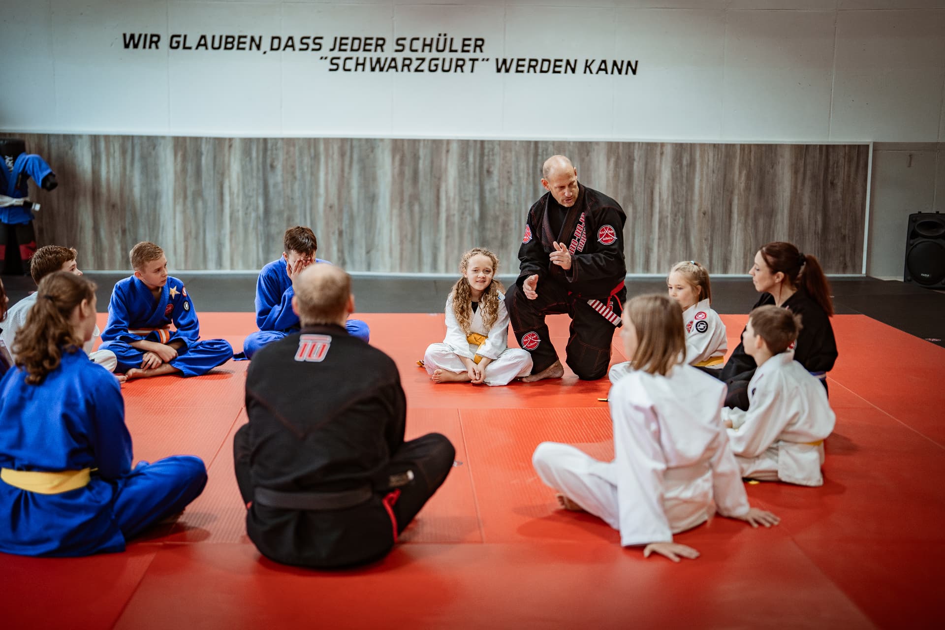 Trainer sitzt mit Kindern und Jugendlichen im Kreis auf der roten Matte im SAO-DOJO Geldern