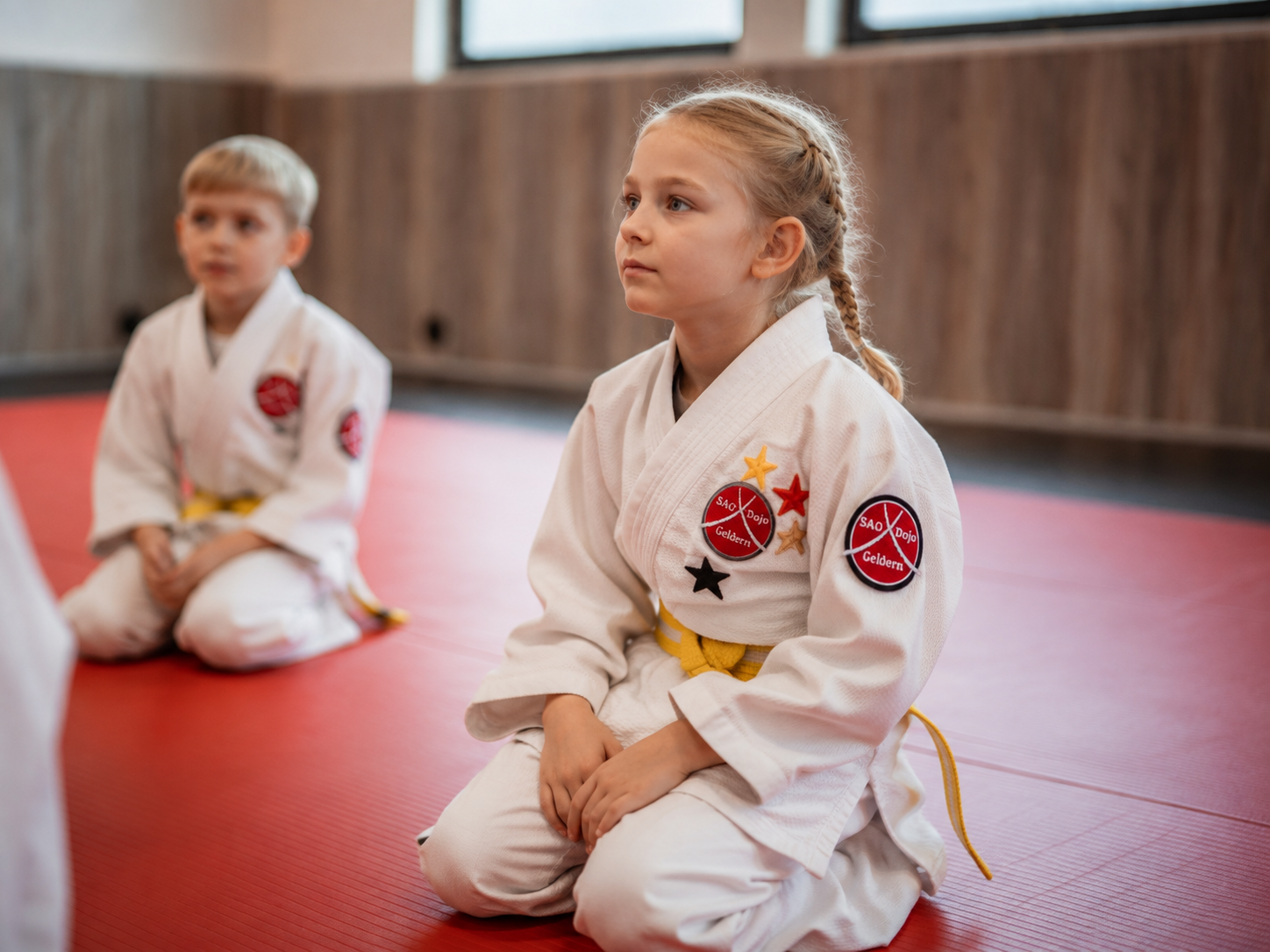 Kind im weißen Anzug sitzt aufmerksam auf der roten Matte im SAO-DOJO Geldern